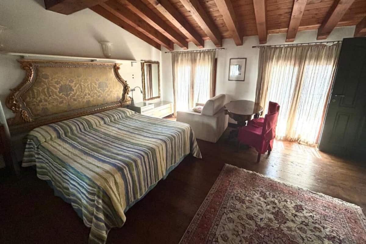 A bedroom with a large bed, a wooden ceiling, a small table with a chair, and a window with curtains.
