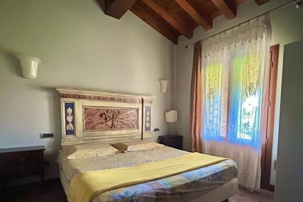 A bedroom with a wooden ceiling, a bed with a yellow blanket, a patterned rug, and a window with curtains.
