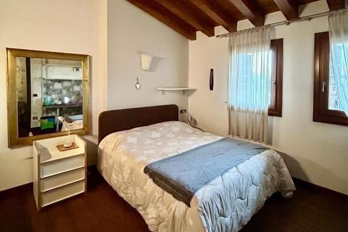 A bedroom with a wooden ceiling, a bed with a floral bedspread, a bedside table, a mirror, and a window with curtains.