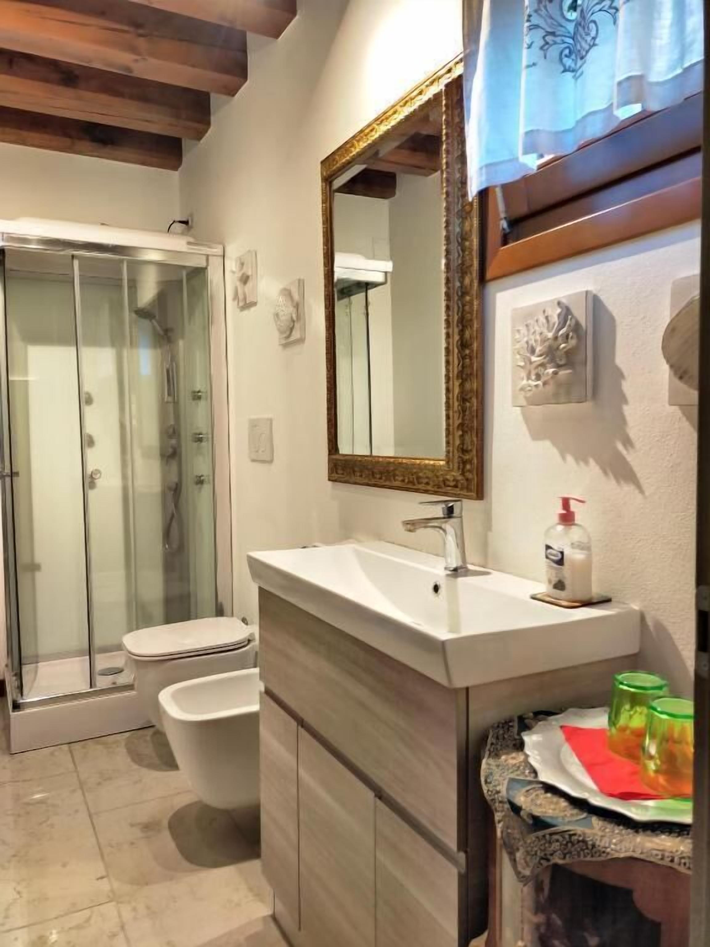 Une salle de bain avec une douche à parois de verre, un lavabo blanc avec un miroir doré et des toilettes.