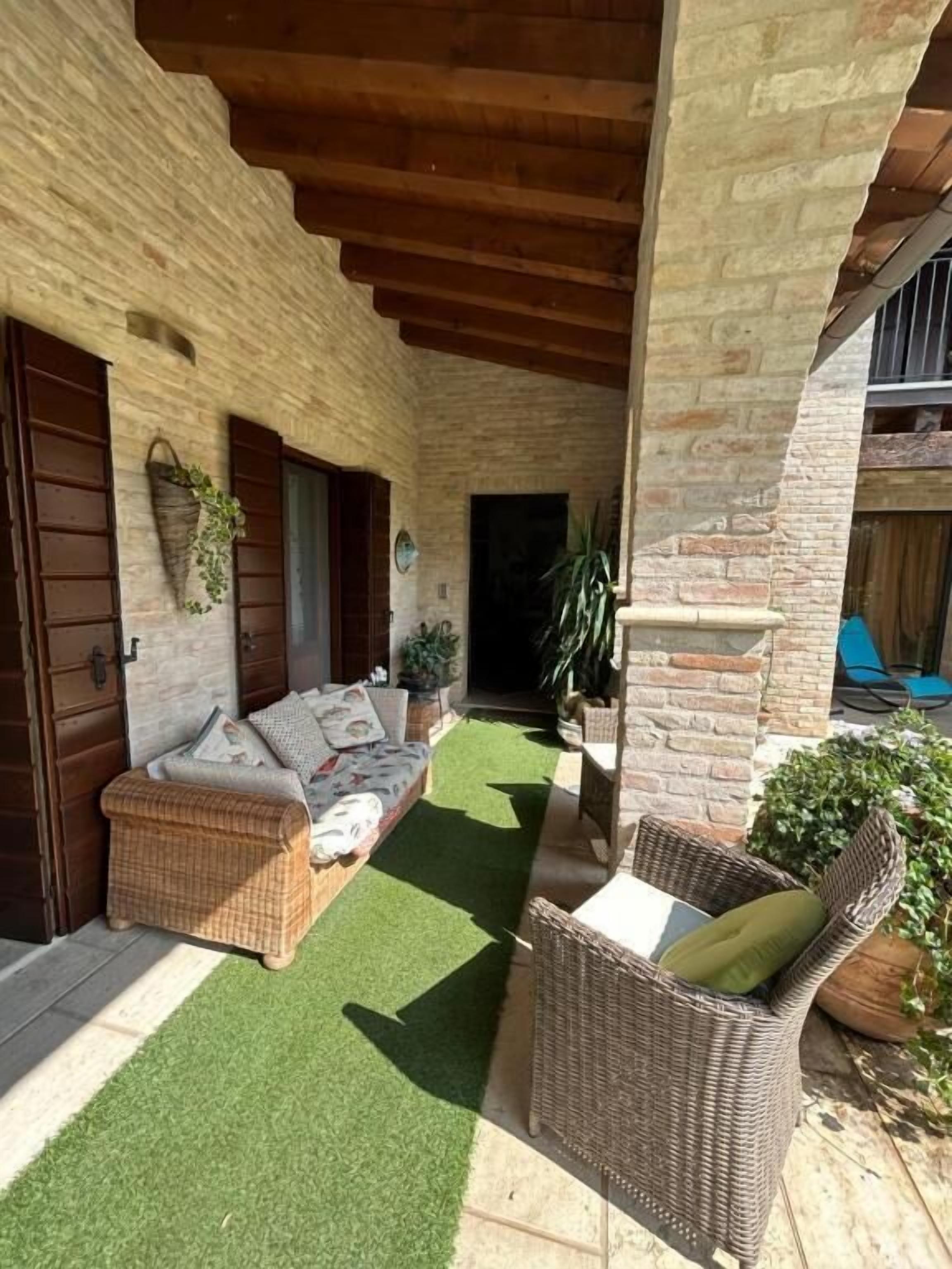 Un'area salotto esterna coperta con divano e poltrone in vimini, un tavolino e piante in vaso.