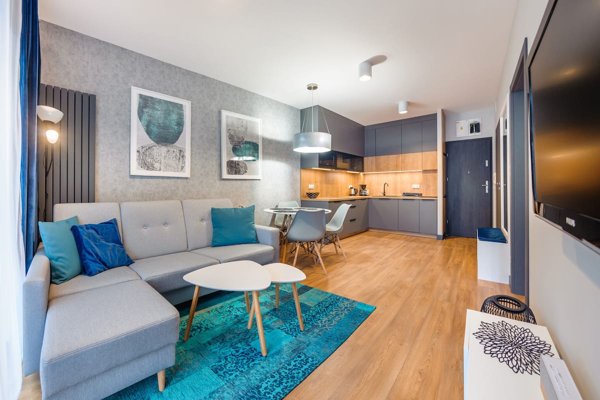 Een moderne woonkamer met een grijze modulaire bank, een witte salontafel en een blauw gestreept vloerkleed. Er is een keuken met houten kasten en een eettafel met stoelen.