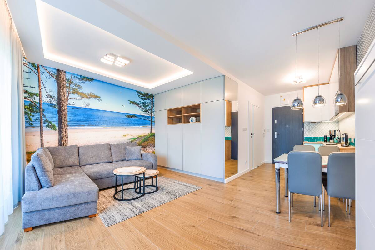 Een moderne woonkamer met een bank, een klein rond tafeltje en een eethoek met stoelen. In de achtergrond is een keuken zichtbaar.