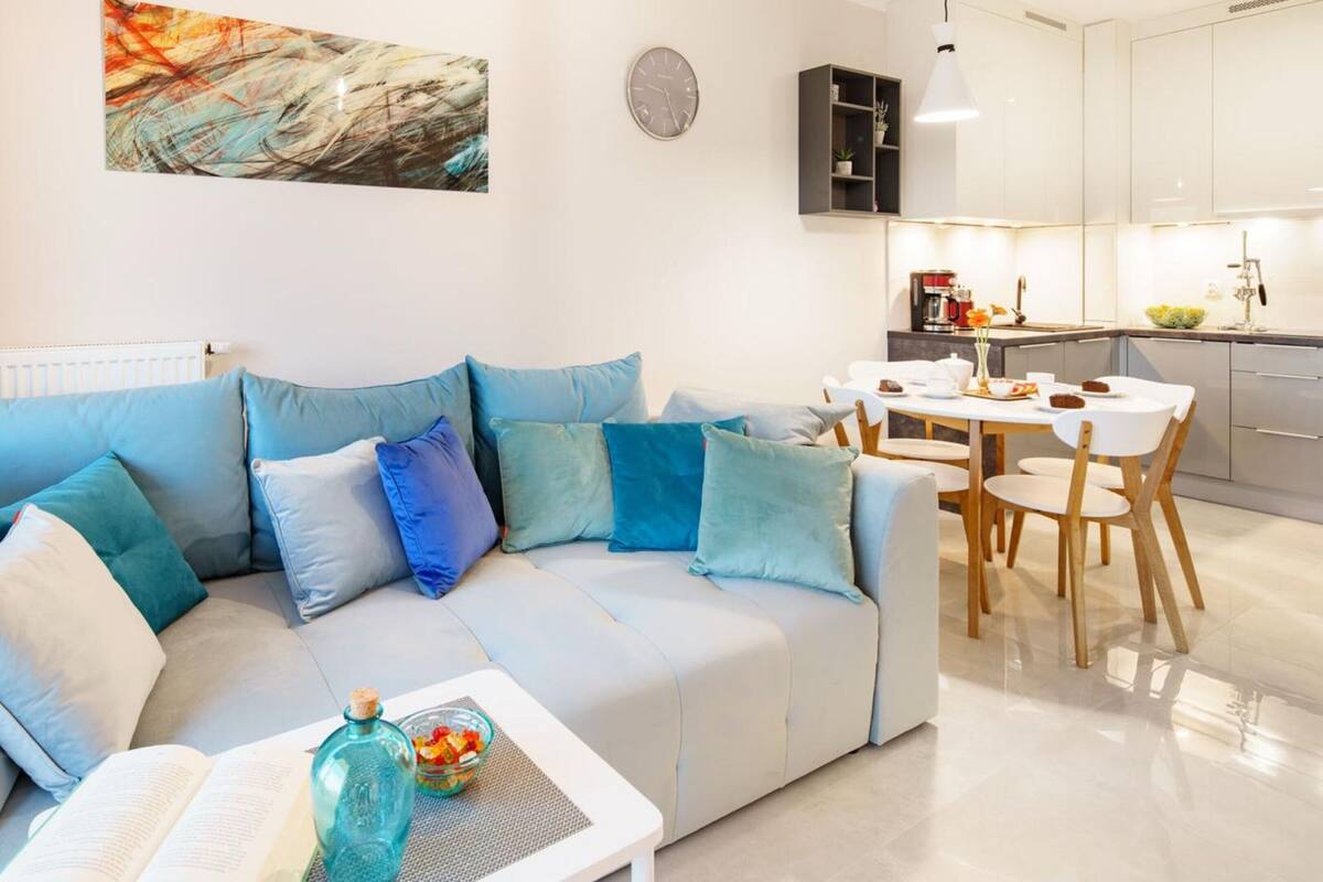 Een moderne woonkamer met een lichtblauwe bank, een bijzettafeltje met een boek en een glazen fles, en een eethoek met een witte tafel en stoelen.