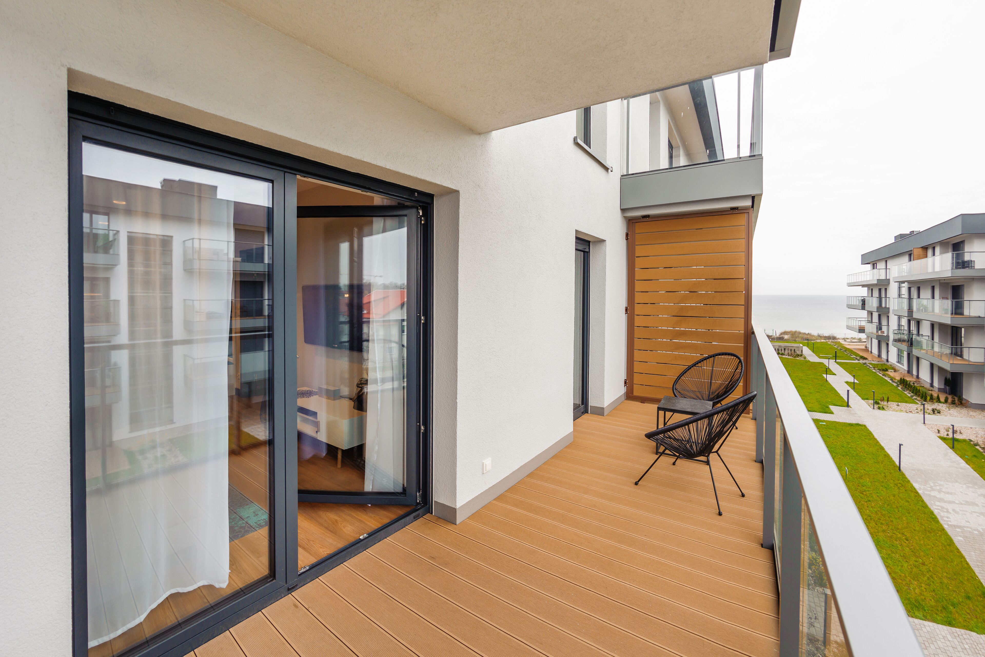 Un balcone con pavimento in legno, una porta scorrevole in vetro e vista su un edificio moderno e il mare.