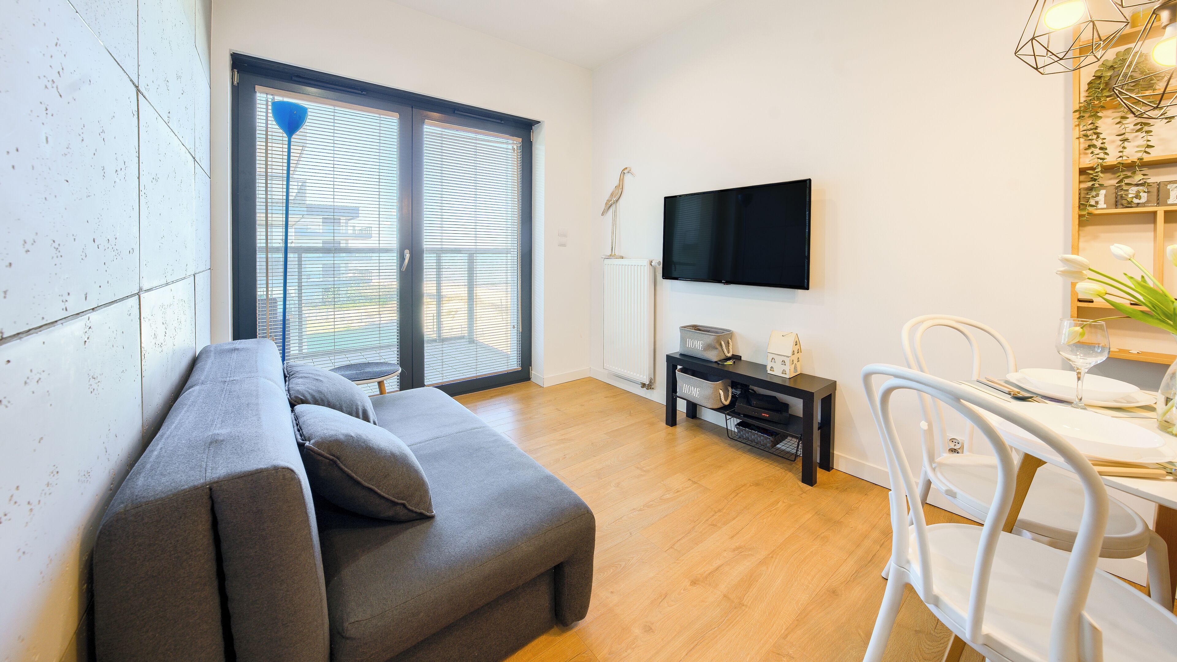 Un salon moderne avec un canapé gris, une table à manger avec des chaises, un téléviseur fixé au mur et une fenêtre avec des stores.