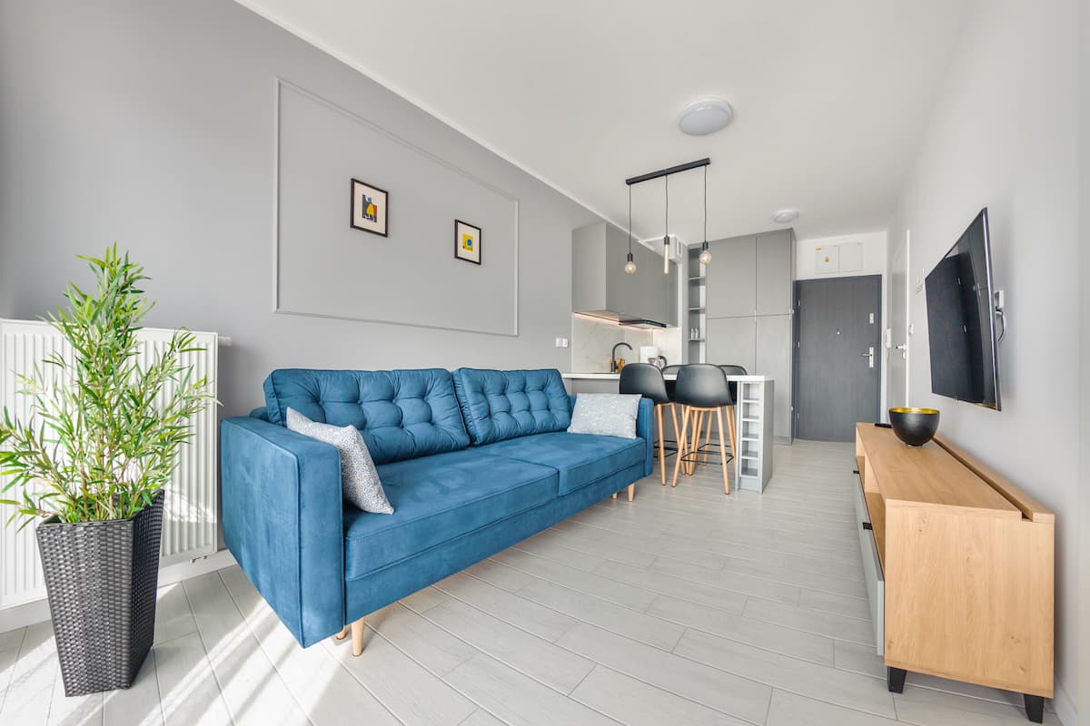 Een moderne woonkamer met een blauwe bank, een eethoek met stoelen, een houten dressoir en een aan de muur bevestigde televisie.