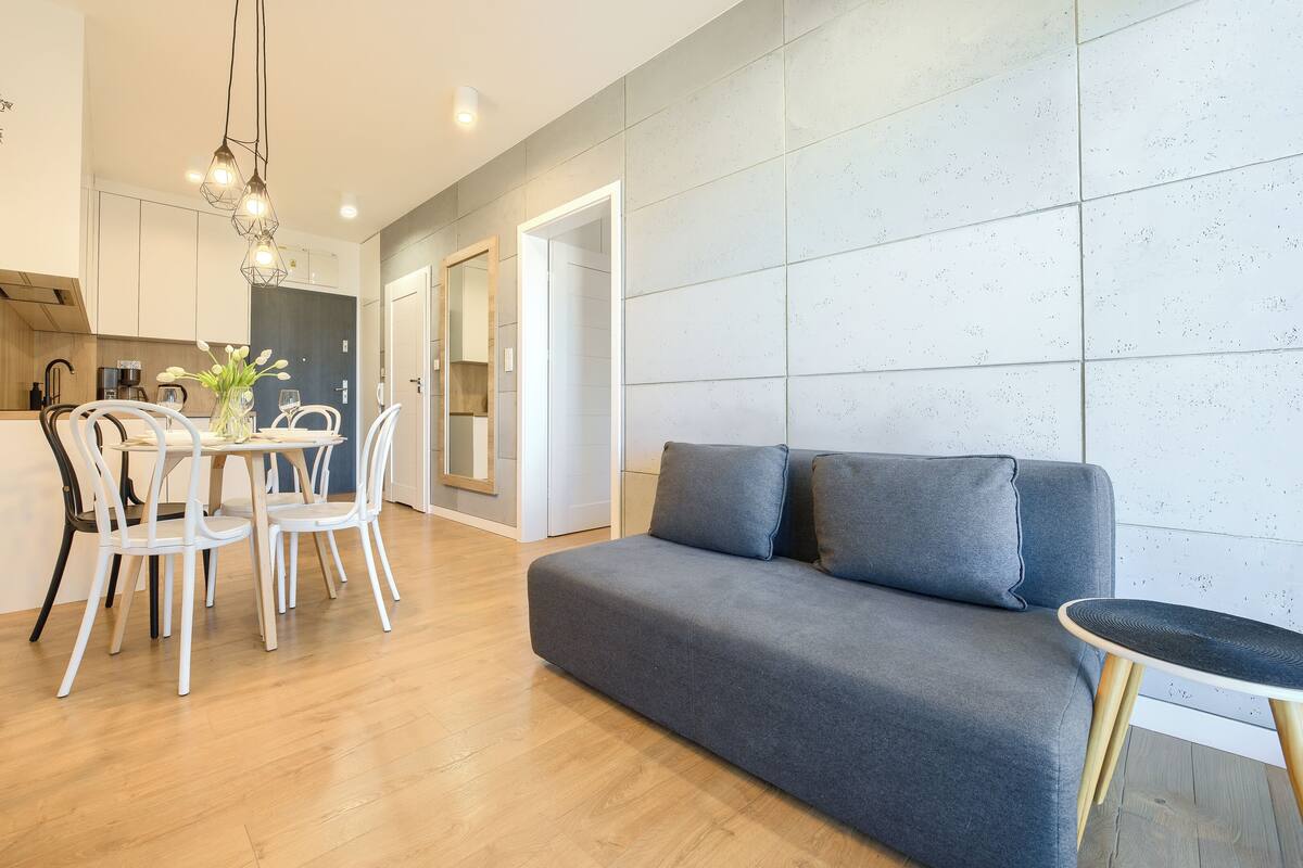 Een moderne woonkamer met een grijze bank, witte stoelen, een eettafel en een keuken met houten kastjes en een hanglamp.
