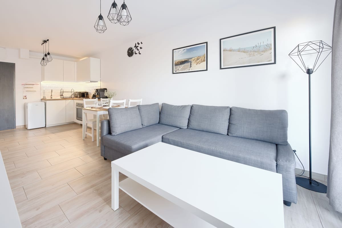 Een moderne woonkamer met een grijze hoekbank, een witte salontafel en een keuken op de achtergrond.
