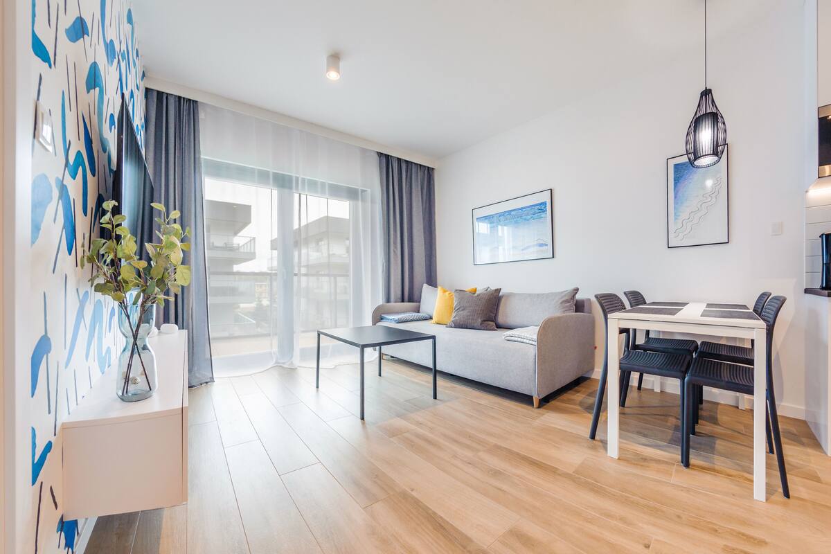 Een moderne woonkamer met een bank, salontafel en een eettafel met stoelen. Er is een groot raam met gordijnen, een kunstwerk aan de muur en een hanglamp.