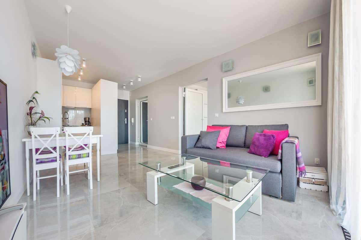 Een moderne woonkamer met een glazen salontafel, een grijze bank met roze kussens, en een eethoek met witte stoelen.