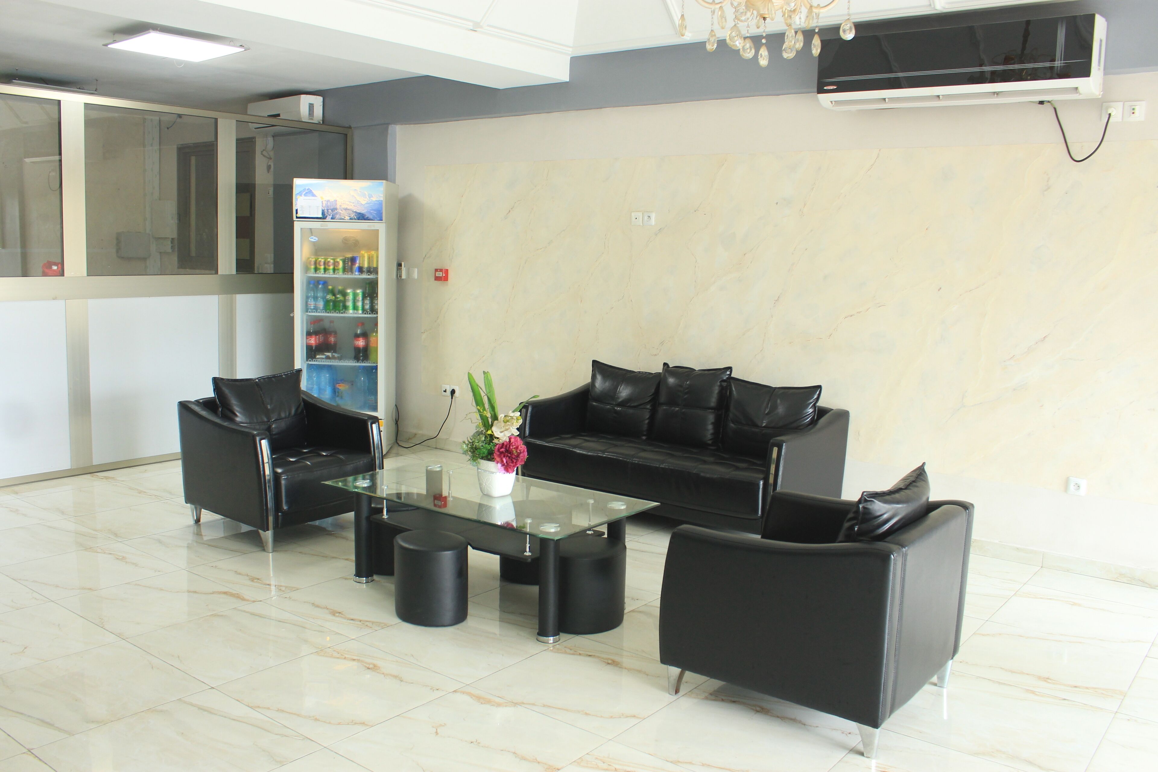 A modern lobby with a glass coffee table, black leather sofas, a refrigerator stocked with beverages, and a marble floor.