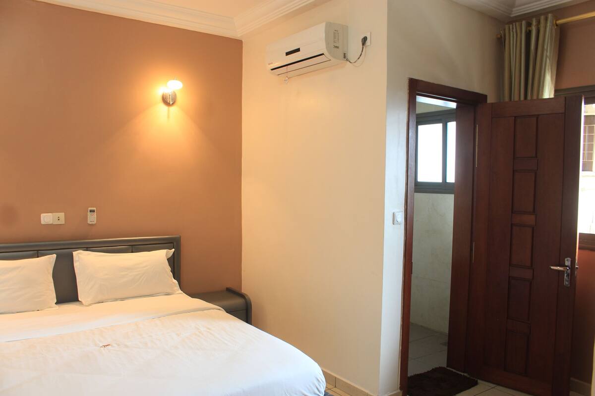 A hotel room with a bed, bedside table, wall-mounted air conditioning unit, and a door leading to another room.