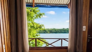 Un lit simple avec un oreiller blanc et une serviette, des murs en bois, une vue sur un lac et des rideaux.