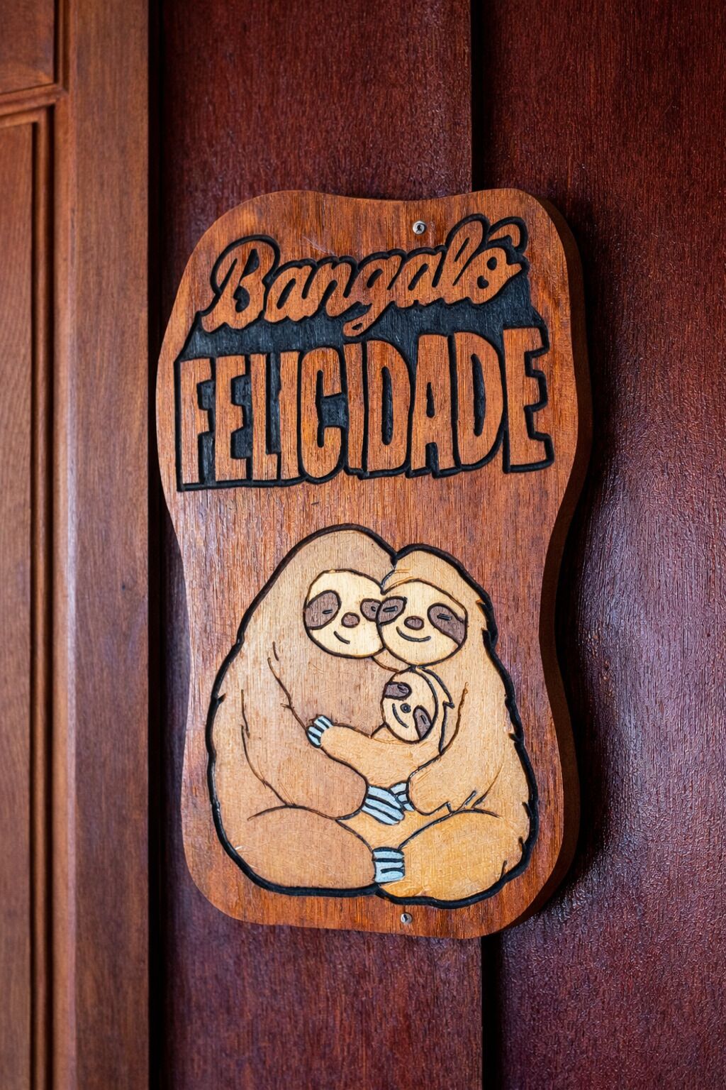 A wooden plaque with a sloth illustration and the text 'Bangáes FELICIDADE'.