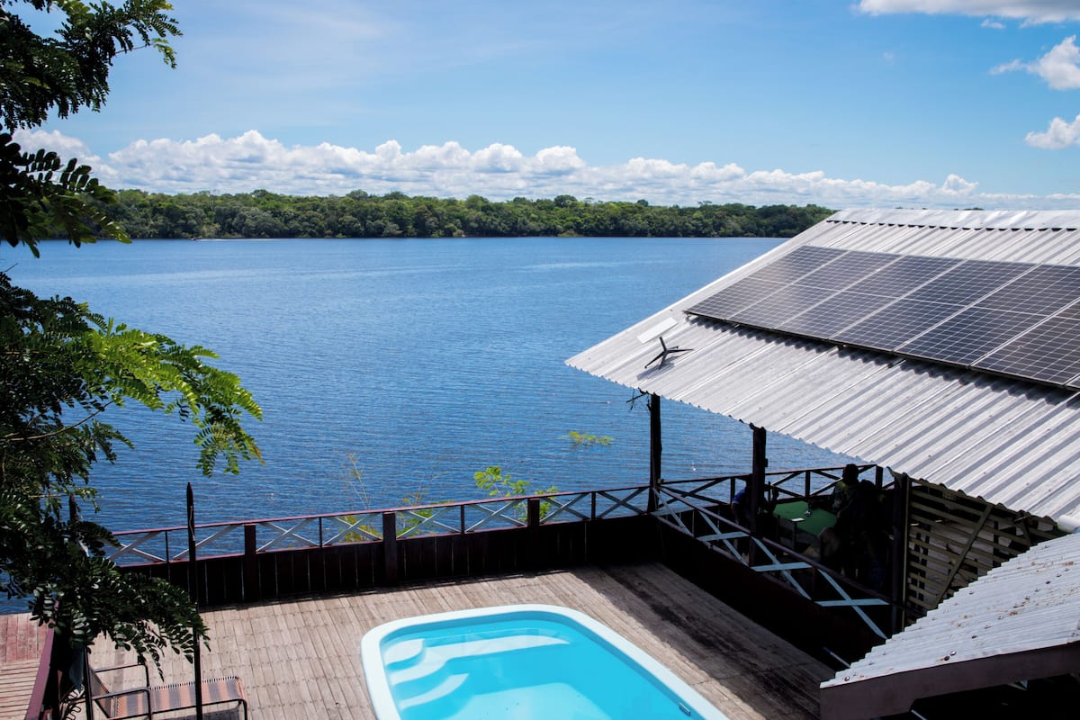 Un espace piscine avec vue sur un lac, une structure couverte équipée de panneaux solaires et une terrasse en bois.