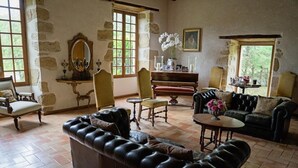 Una habitación con un amplio sofá de cuero, un piano, una mesa de comedor y una silla. Se ven vistas de follaje a través de las ventanas.