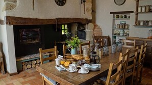 Un comedor rústico con una mesa de madera dispuesta para el desayuno, rodeada de sillas. Se observa una chimenea de ladrillo con chimenea al fondo.