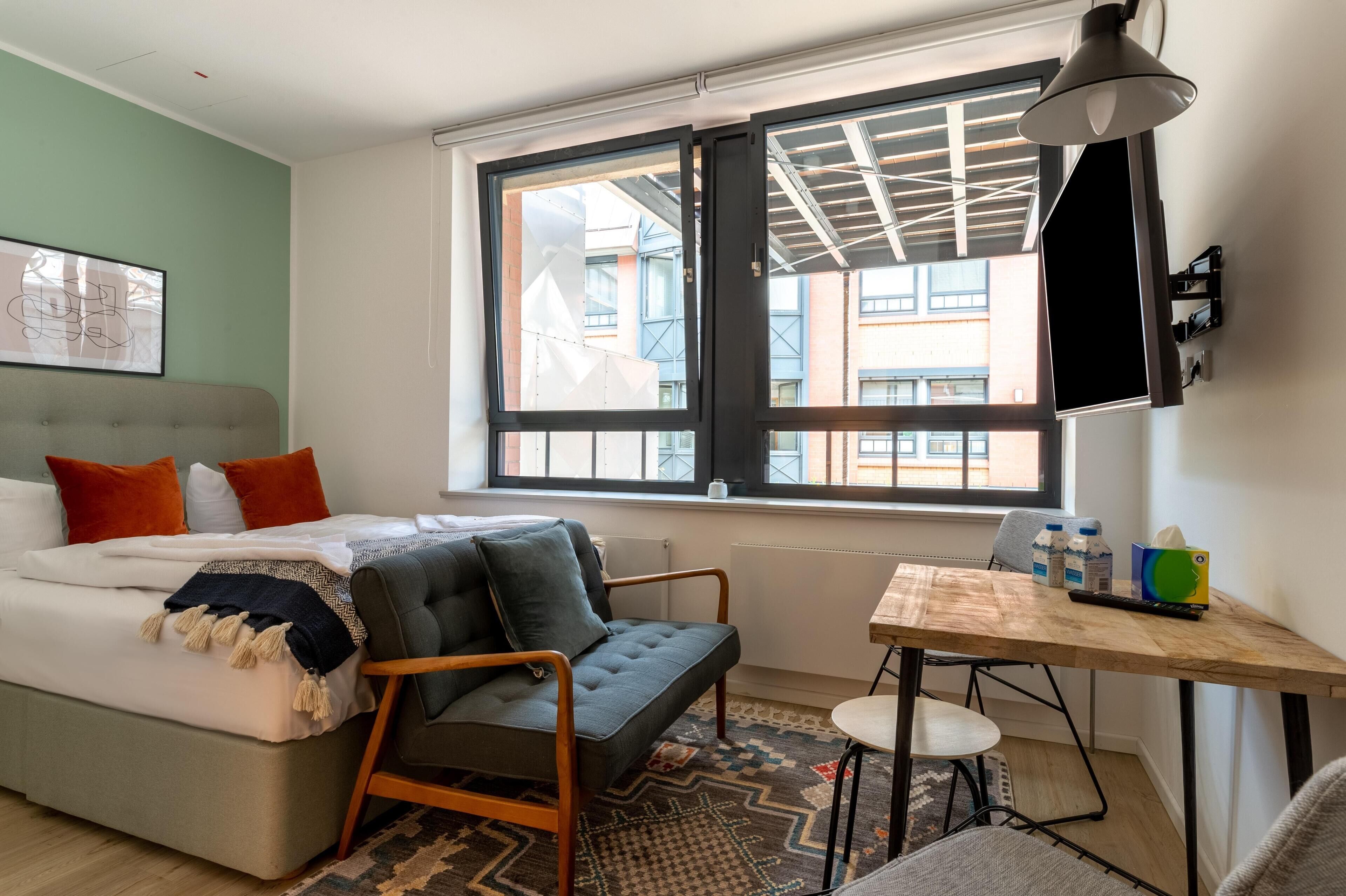 Ein modernes Schlafzimmer mit Bett, Sofa, Esstisch und einem großen Fenster mit Blick auf Gebäude.