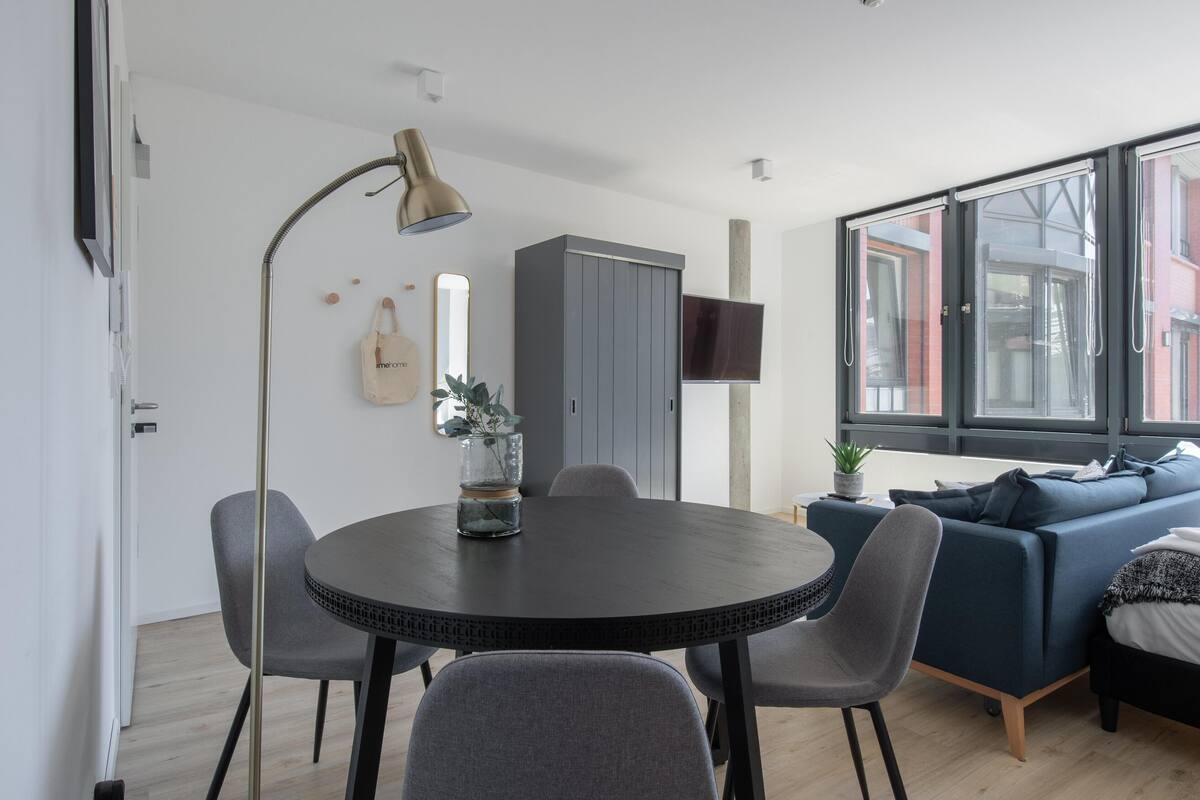Una sala de estar moderna con una mesa de comedor redonda, sillas grises, un sofá azul y un televisor instalado en la pared.