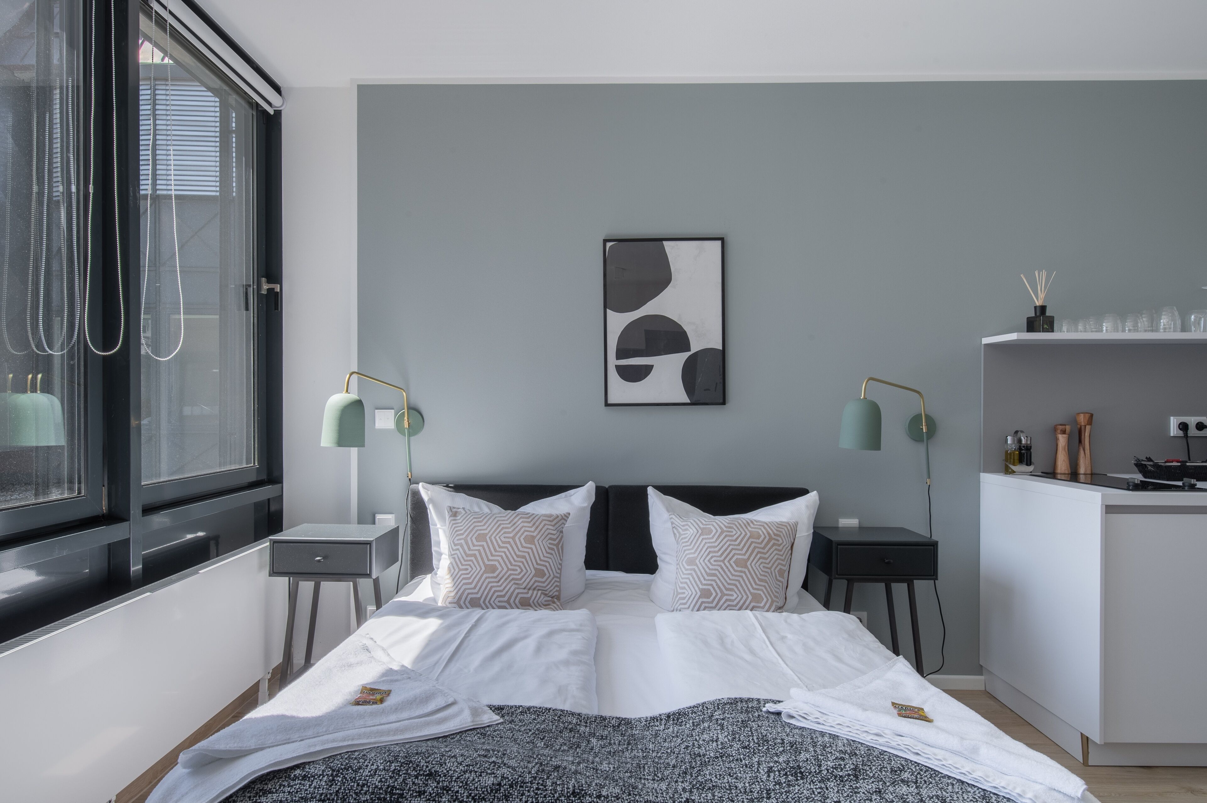 A modern bedroom with a large bed, two bedside tables, a small desk, and a built-in kitchen area.