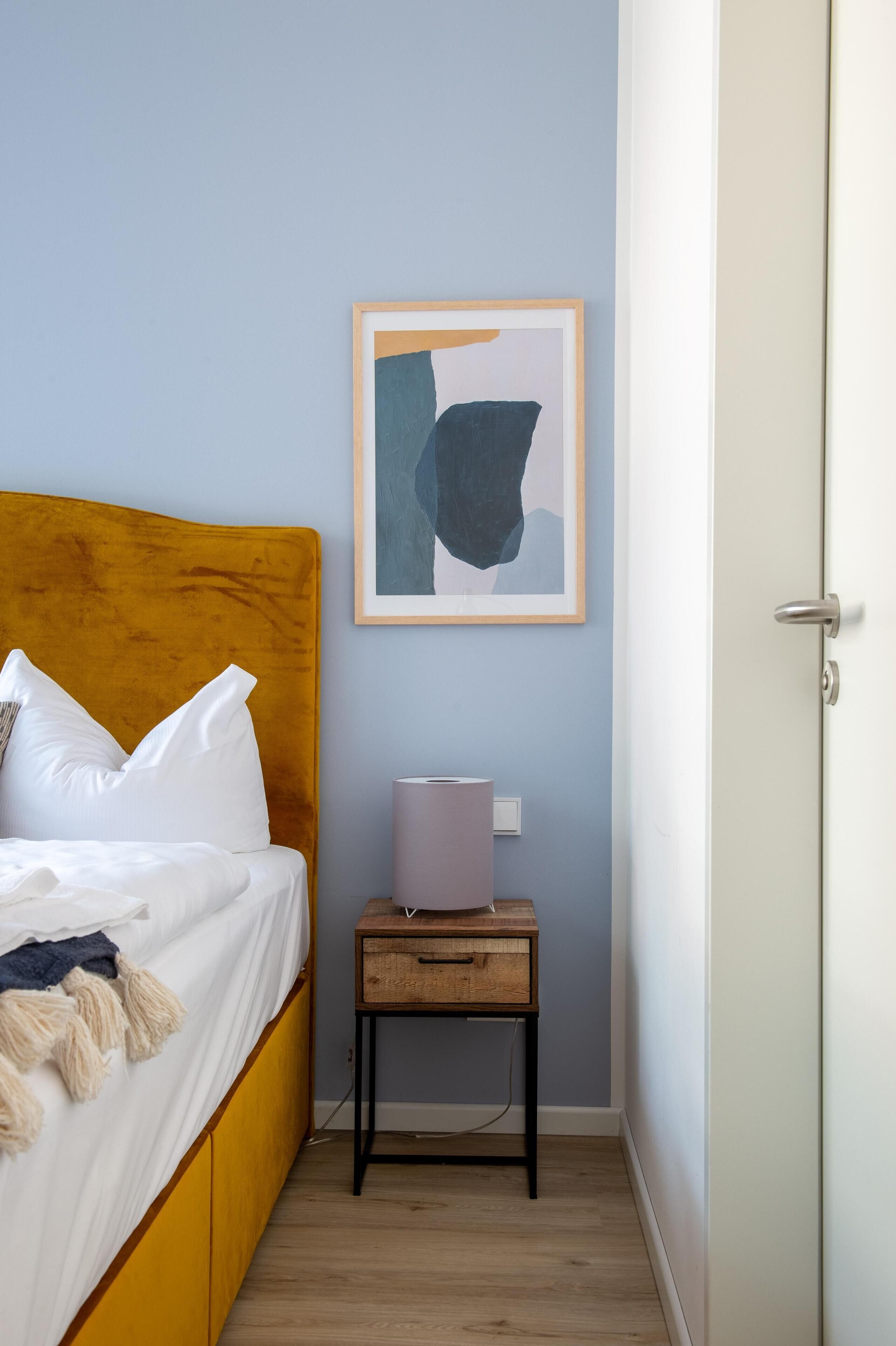 A bedroom with a yellow headboard, white bedding, a bedside table with a lamp, and a framed abstract painting on the wall.