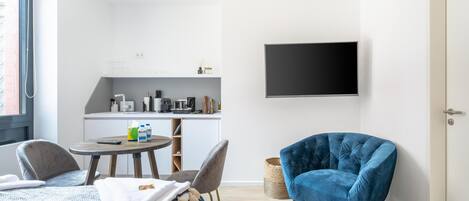 Un espacio habitable compacto con cama, un sillón azul, una mesa de comedor con sillas, una zona de cocina y un televisor de pantalla plana montado en la pared.