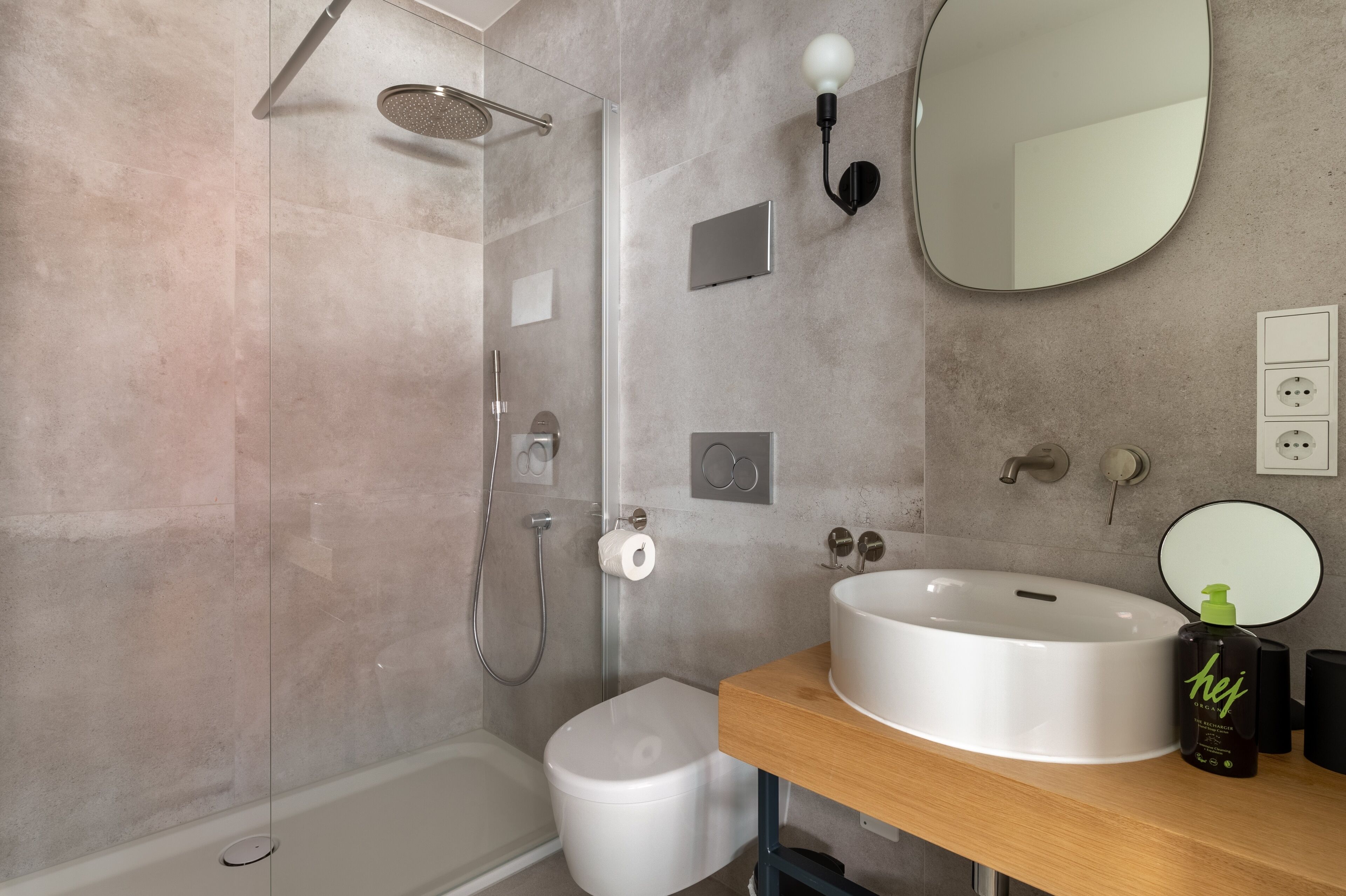 Un baño moderno con ducha de vidrio, inodoro blanco, un espejo redondo grande y un tocador de madera con lavamanos.