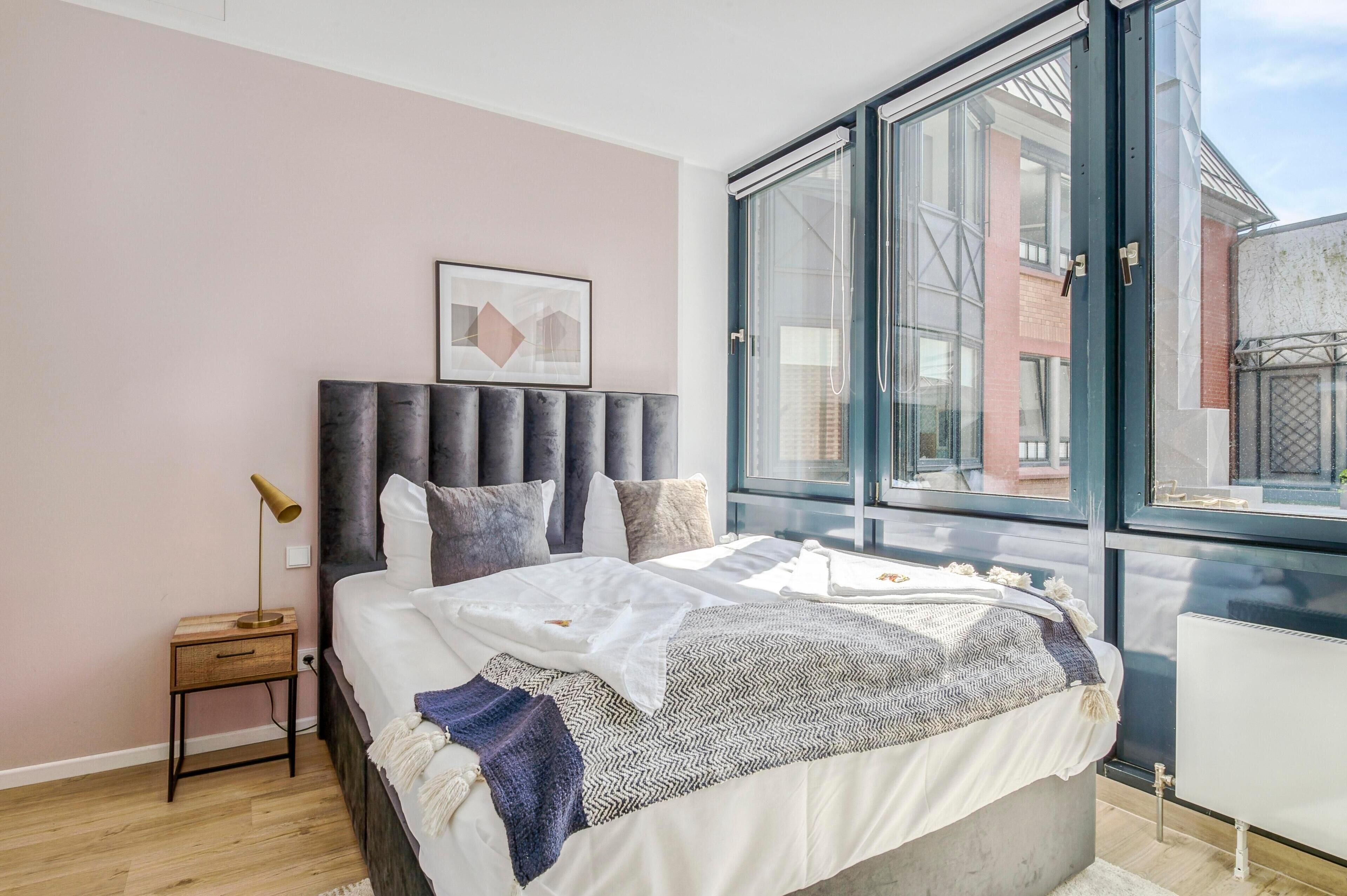 A modern bedroom with a large window, a bed with a grey headboard, and a small bedside table with a lamp.