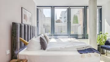 A modern bedroom with a large bed, a bedside table with a lamp, a framed picture on the wall, and a view of the outside through large windows.
