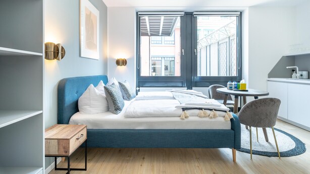 Un dormitorio moderno con cama, mesitas de noche, una silla, una pequeña mesa y vistas a través de un ventanal.