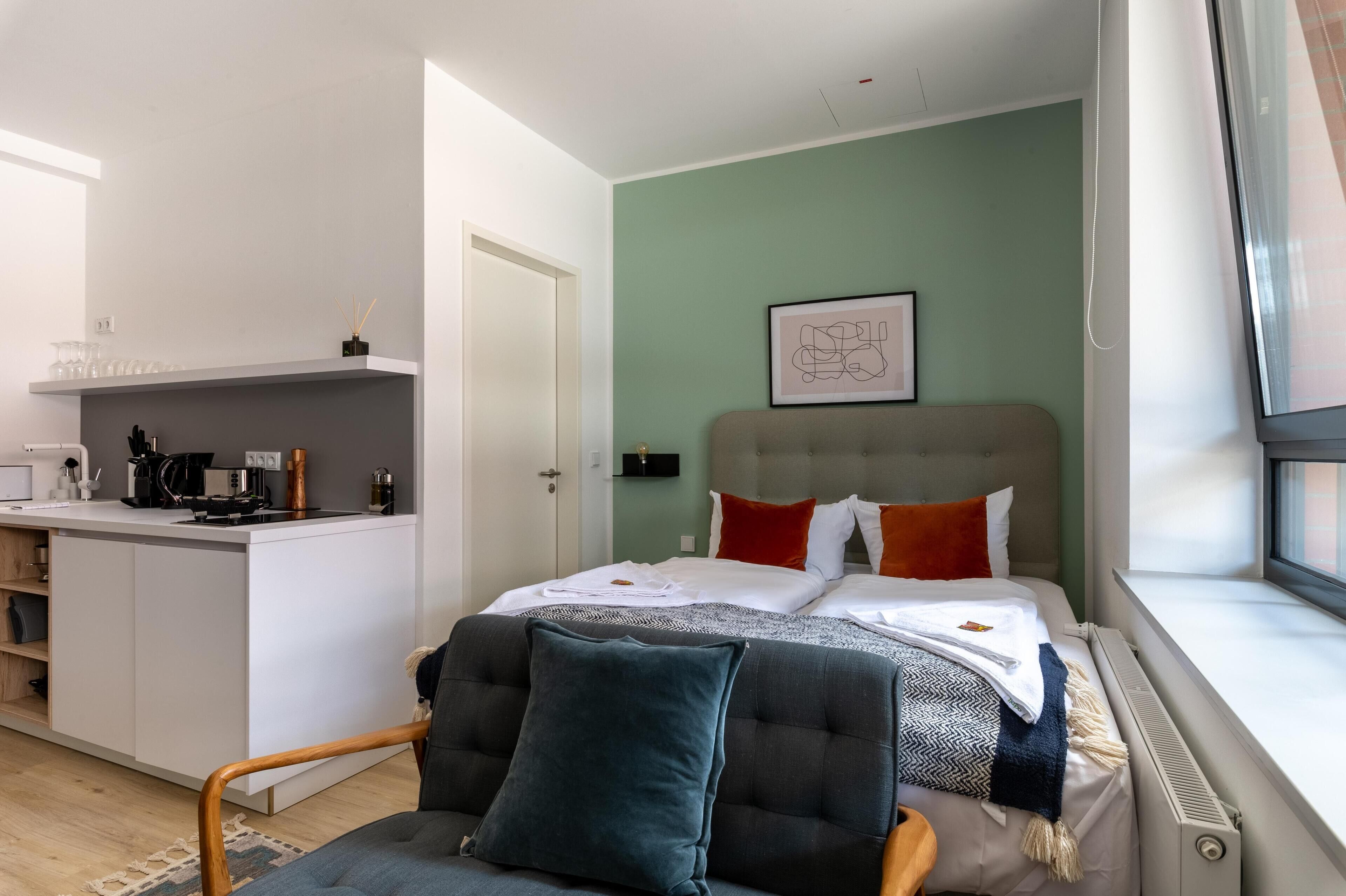 Habitación de hotel moderna con kitchenette, cama con varias almohadas, ventana con vistas y una obra de arte enmarcada en la pared.