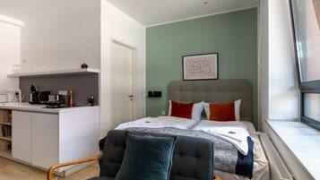 A modern hotel room with a kitchenette, a bed with multiple pillows, a window with a view, and a framed artwork on the wall.