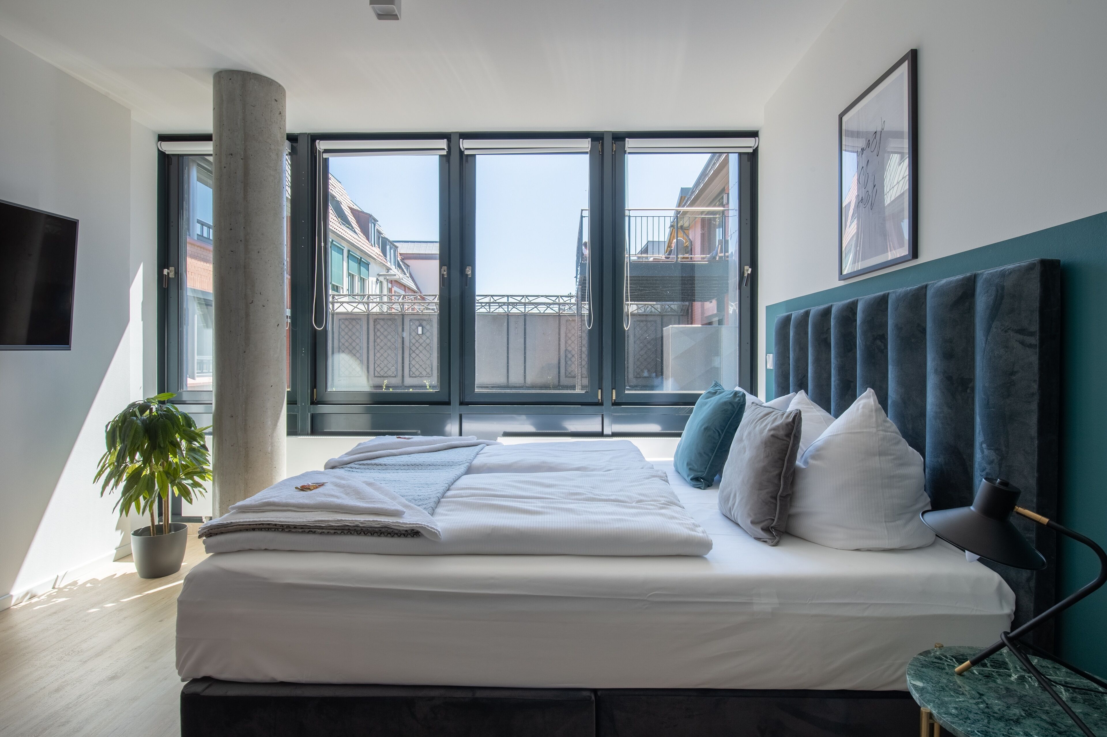 Une chambre moderne avec un grand lit, une plante en pot, un bureau avec une lampe et une vue sur les immeubles à travers de grandes fenêtres.