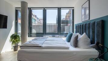 Een moderne slaapkamer met een groot bed, een potplant, een bureau met een lamp, en een uitzicht op gebouwen door grote ramen.