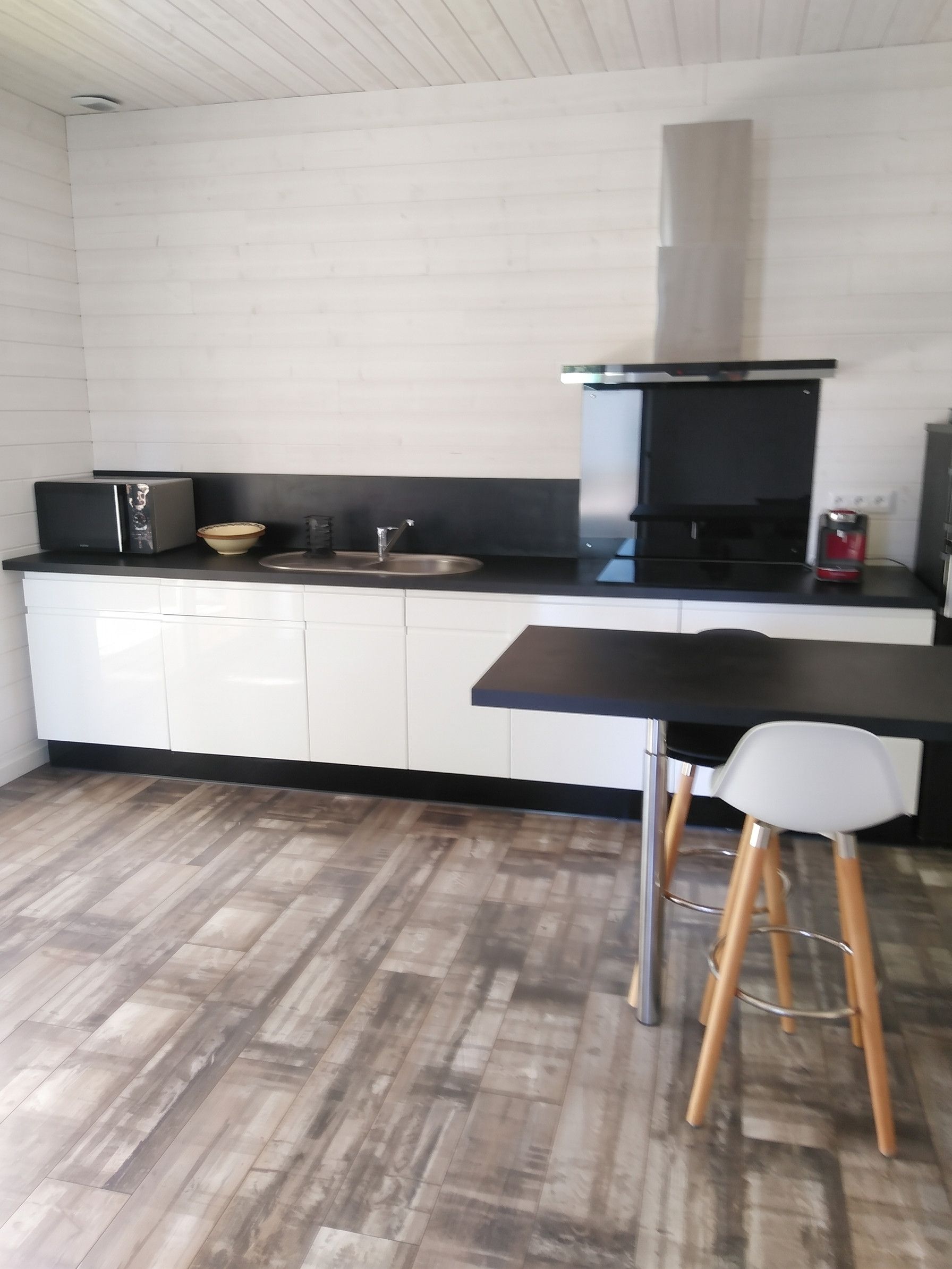 Ett modernt kök med en svart bänkskiva, vita skåp, en diskho i rostfritt stål och en svart spis. Det finns en barstol med en vit sits och ben av trä.