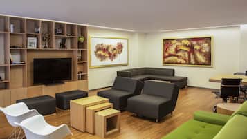 Un salon moderne avec un téléviseur à écran plat, des canapés gris, des fauteuils blancs et verts, des tables d’appoint en bois et des tableaux abstraits accrochés aux murs.