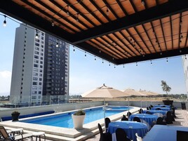 Un área de piscina en la azotea con mesas y sillas, un edificio grande al fondo y luces colgantes.