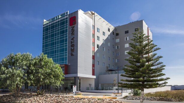 A modern hotel with a prominent glass facade, a mix of white and gray exterior, and a red logo. - Fiesta Inn Celaya Galerías (Celaya)