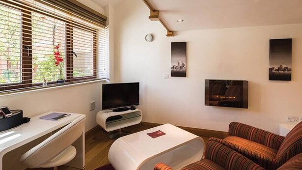 A modern living room with a flat-screen TV, a sofa, a desk with a chair, and a window with blinds.