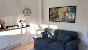 A modern kitchen with white cabinets, a sink, and a clock. There is a gray sofa with striped pillows and a colorful abstract painting on the wall.