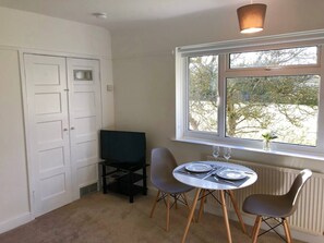 Quarto pequeno com uma mesa de jantar, duas cadeiras, uma televisão e uma janela com vista para árvores.
