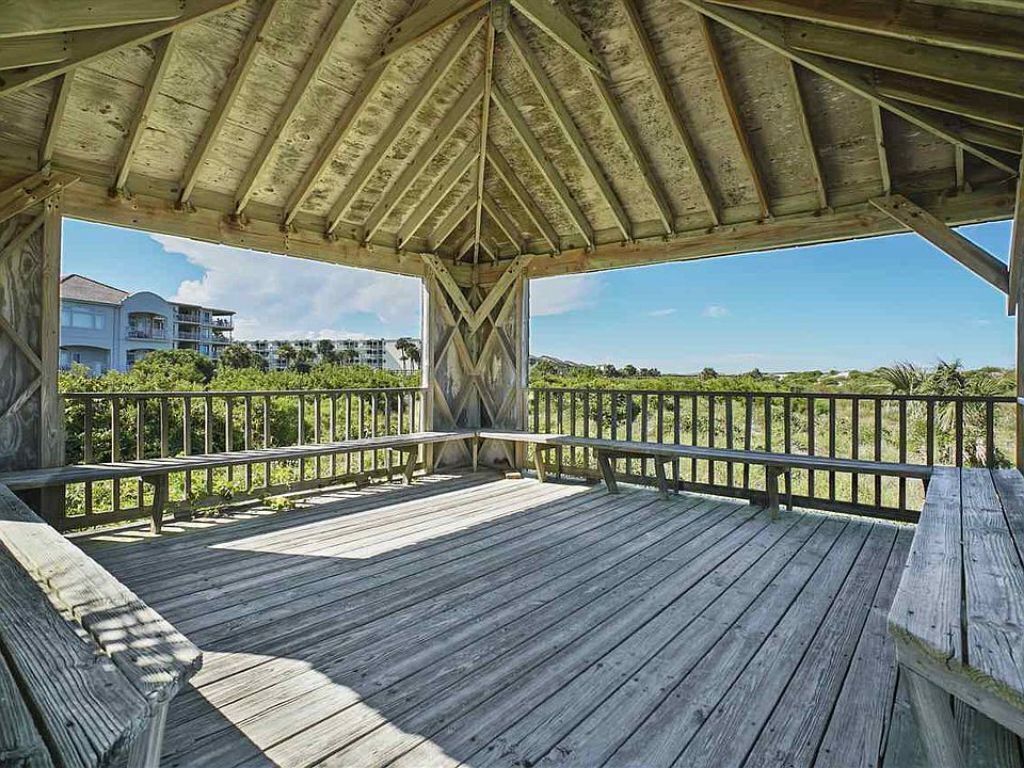 Una terraza de madera con barandilla metálica, con vista a una playa y edificios al fondo.