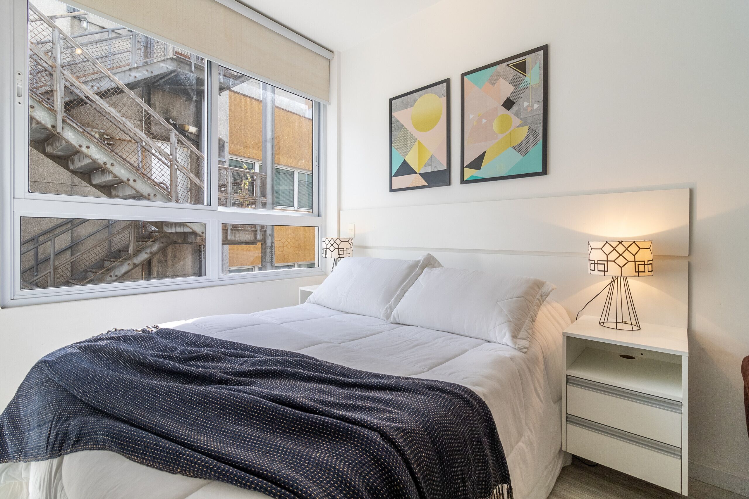 A bedroom with a large window, a bed with white linens, a dark patterned throw blanket, two wall-mounted lamps, and abstract artwork.