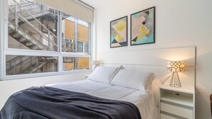 A bedroom with a large window, a bed with white linens, a dark patterned throw blanket, two wall-mounted lamps, and abstract artwork. - AV1209 - Studio 4p/ 1q (São Paulo)
