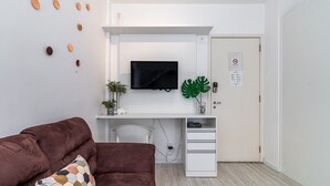 A modern room with a white desk, a mounted TV, a door with a security panel, and a brown sofa with a patterned pillow. - AV1209 - Studio 4p/ 1q (São Paulo)