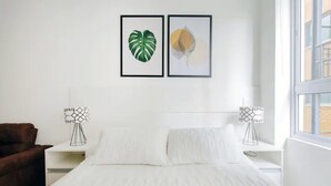 Camera da letto con un letto, pareti bianche, due quadri appesi, una sedia marrone e due lampade sui comodini.