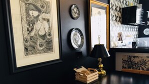 A dark-themed room with a wooden chair, a table with a lamp, and framed artwork on the wall.