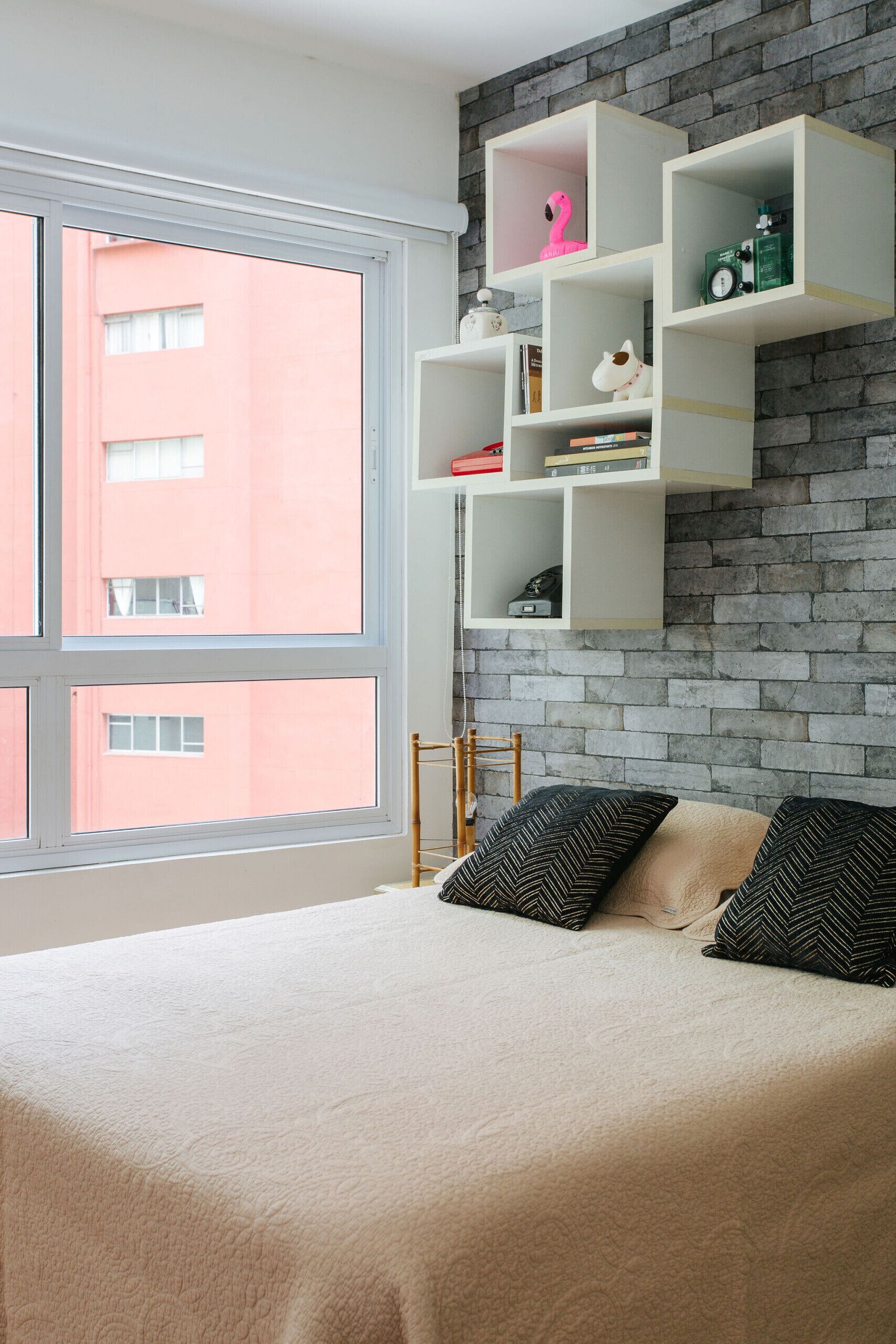 Un dormitorio con un gran ventanal, una cama con almohadas estampadas y estantes blancos flotantes contra una pared de ladrillo.