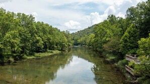 Ein Fluss, umgeben von dichtem Grün, mit einer Holzbrücke auf der rechten Seite.